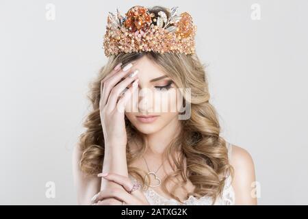 Sad Beautiful blonde girl in a golden crown on white grey background, eyes closed looking down with grief hand on shoulder pink ring on it Stock Photo