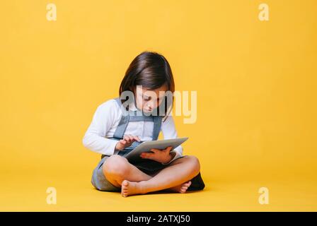A caucasian girl in a gray dress and white shirt uses and plays with a tablet on a yellow background Stock Photo