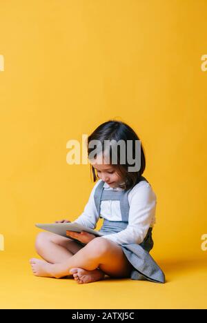 A caucasian girl in a gray dress and white shirt uses and plays with a tablet on a yellow background Stock Photo