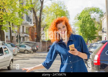 Taxi from app. Pretty happy woman holding mobile phone, hitch hiking in the city street on a sunny day. One single person. Stop Sign, Lifestyle. Stock Photo