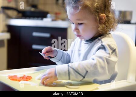 Kid cuts carrots and potatoes in kitchen. Cute two-year little girl helps the mother to cook. Independent little child learns to cook food. Beautiful Stock Photo