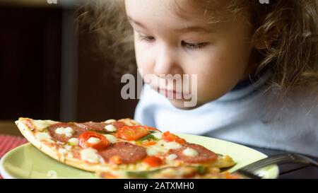 Child looks at pizza on table. Three years old kid is going to eat by himself. Little girl and food on plate. Feeding cute baby at home. Portrait of n Stock Photo