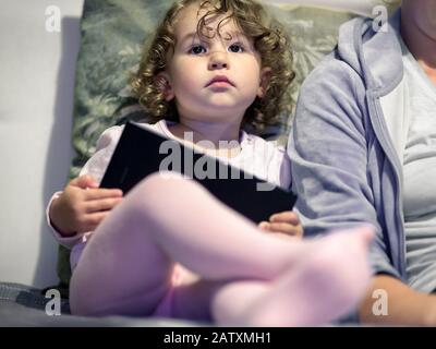 Baby girl sits near her mother and uses a digital tablet. Two-year child looks electronic device and TV at home. Cute baby plays with the gadget. Todd Stock Photo