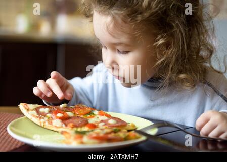 Child looks at pizza on table. Three years old kid is going to eat by himself. Little girl wants to take a piece of food from plate. Feeding cute baby Stock Photo