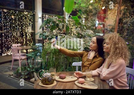 Asian young woman holding her mobile phone and making selfie portrait with her friend while they drinking tea in cafe Stock Photo