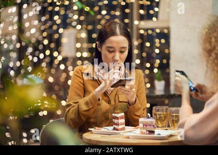 Asian pretty woman using her mobile phone while eating dessert during a meeting with her friend in cafe Stock Photo