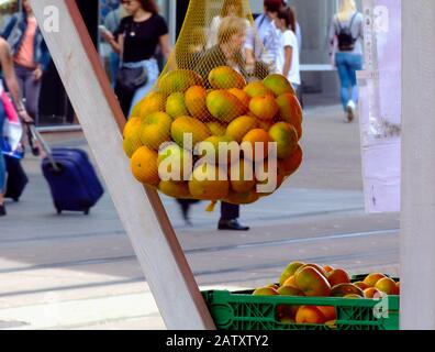 fruit vendor stand. orange, yellow and green clementines. vinyl net sack suspended from wood frame. blurry urban background with tourist pedestrians Stock Photo