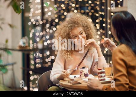 Young beautiful woman with curly hair eating dessert and enjoying the time with her friend in cafe Stock Photo