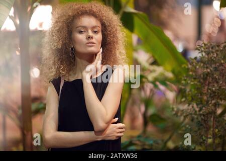 Portrait of beautiful young woman with blonde curly hair in black dress posing at camera Stock Photo