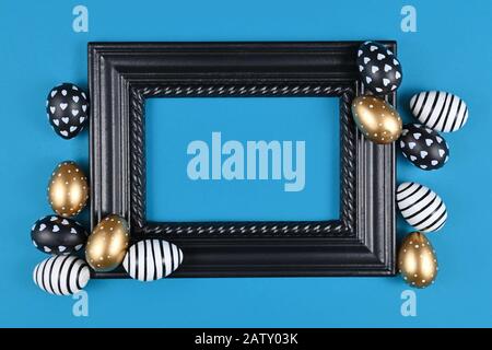 Easter design with blank black elegant picture frame surrounded by modern golden, black and white easter eggs on blue background Stock Photo