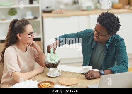 African young man sitting at the table and pouring coffee into the cup of his girlfriend while they sitting in the kitchen Stock Photo