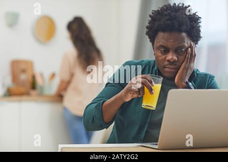 African young guy drinking juice and using laptop computer at the table in the kitchen Stock Photo