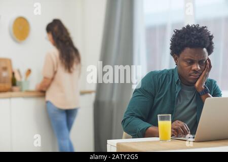 African serious man sitting at the table with glass of juice and working on laptop with his girlfriend cooking in the background in the kitchen Stock Photo