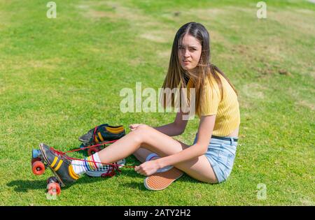 Pretty teenage girl in yellow top sits on grass putting retro style roller skates on getting ready to go skating. Stock Photo