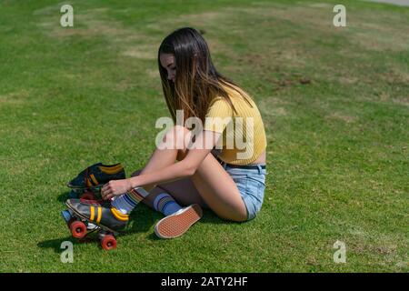 Pretty teenage girl in yellow top sits on grass putting retro style roller skates on getting ready to go skating. Stock Photo