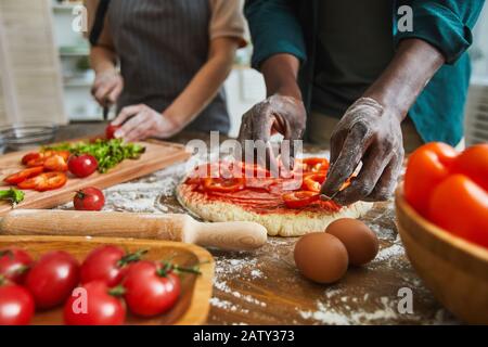 Close-up of African man putting pepper on dough for future pizza with woman cutting vegetables in the background Stock Photo