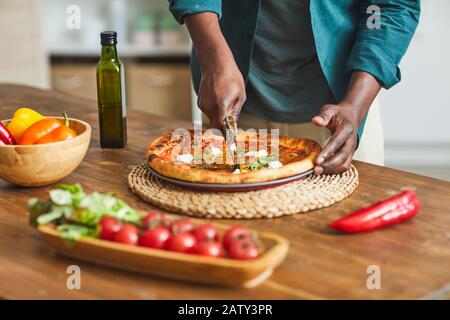 Close-up of African young man standing near the wooden table and cutting pizza with cheese before eating it Stock Photo