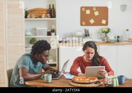 Young multiethnic couple sitting at the table and busy with their gadgets during lunch in the kitchen at home Stock Photo