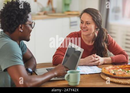 Young woman smiling and talking to African man while they sitting at the table with laptop and documents and planning work in team Stock Photo