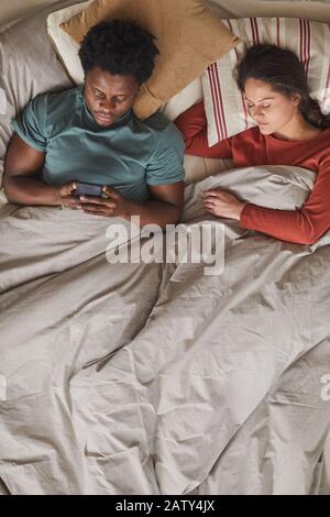 African young man lying on bed with his girlfriend and reading online using his mobile phone in the bedroom Stock Photo