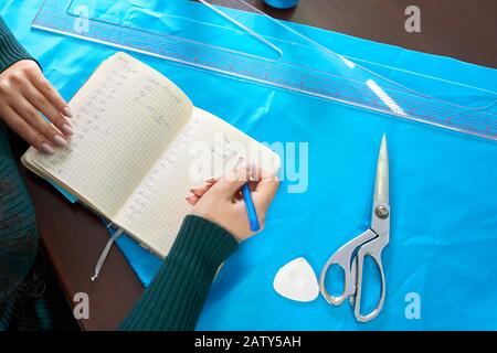 beautiful seamstress with long hair writes in a notebook. the tailor creates a collection of outfits. young woman designer clothes notes ideas in note Stock Photo