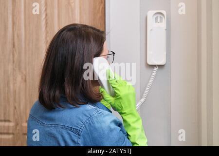 Woman at home near front door talking on intercom, answering the call, holding security phone in hand Stock Photo
