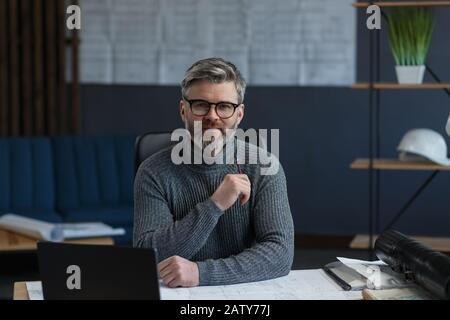 Business portrait of handsome bearded man wearing eyeglasses sitting at workplace. Confident businessman became successful. Interior designer working Stock Photo