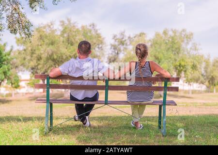 Couple sitting on a wooden bench at a public park with their backs to the camera on an out of focus background. Relax and leisure concept. Stock Photo