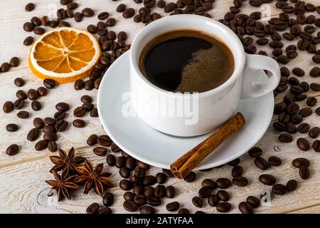Hot coffee in white elegant cup with saucer on a background of coffe beans over a white wooden table. Coffee with cinnamon, anise and dry citrus. Stock Photo