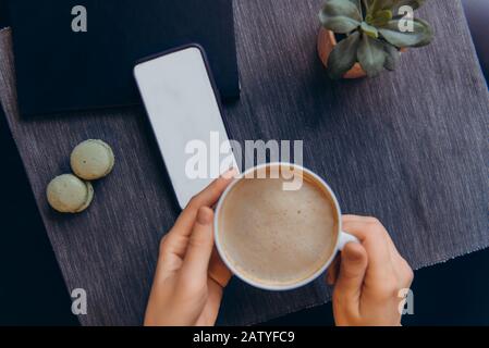 Top view smartphone with white blank screen and blurred female hands holding hot cup of coffee on dark gray background. Workplace with notebook and gr Stock Photo