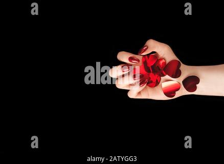 Beautiful female hand holding vibrant red heart-shaped confetti on isolated black background. Backdrop for your design. Creative trendy flat lay with Stock Photo