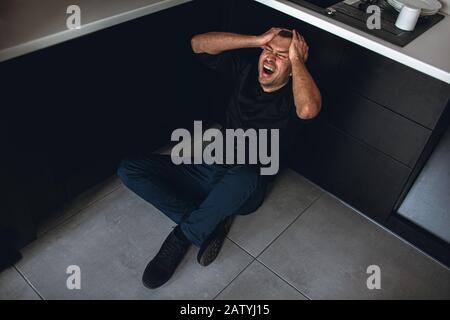 Colorful picture of mad desparate man sitting on floor in kitchen and scream. Hold hands on head. Psychological problems and disorder. Depression and Stock Photo