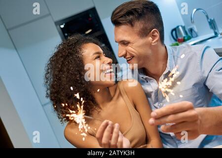 Romantic Date. Young multiethnic couple at home having dinner with sparklers celebrating new year looking at each other happy Stock Photo