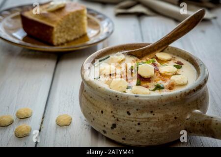 A bowl of homemade creamy potato and bacon soup topped with cheddar cheese, green onions, and oyster crackers with a side of cornbread. Stock Photo