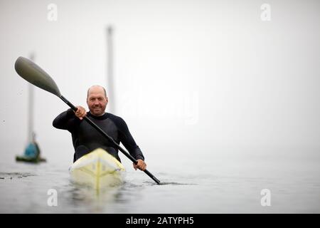 Male kayaker smiles for a portrait as he paddles in the waters of a foggy harbour. Stock Photo