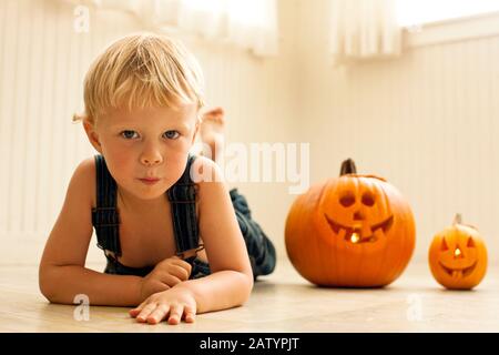 Young boy lies front down on a floor next to a big Jack O'Lantern and a small Jack O'Lantern lit with candles inside as he poses for a portrait. Stock Photo