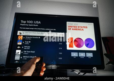 Paris, France - Circa 2019: Man hand holding remote control of Apple TV in front of Panasonic Plasma tv set in living room with top 100 charts USA playlist by Apple Music Stock Photo