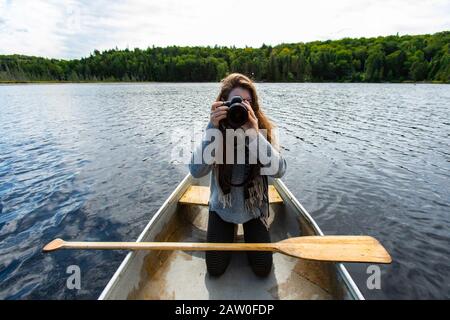 Young female photographer kneeling over knee is canoe clicking pictures with DSLR camera while on lake in Northern Quebec in Canada Stock Photo