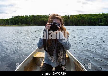 Young female camera person holding DSLR camera and adjusting lens for shoot while riding in canoe on lake in Northern Quebec in Canada Stock Photo