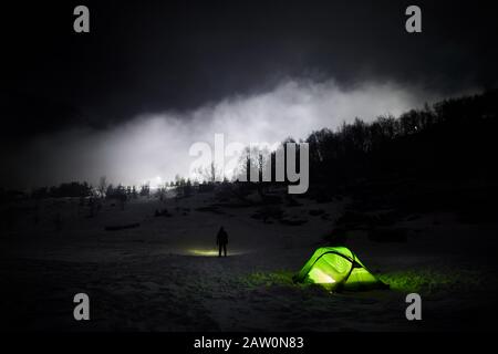 Green glowing tent at frozen lake and silhouette of lonely man against white fog in the mountains at night Stock Photo