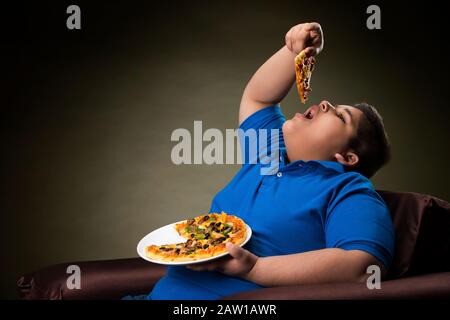 Young boy sitting on the sofa eating a pizza slice. (Obesity) Stock Photo