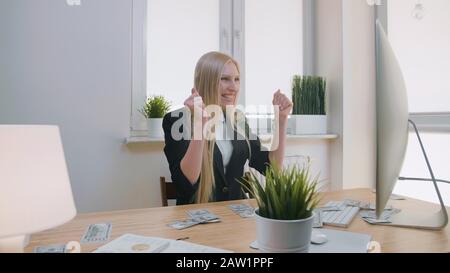Elegant blond female sitting at desk at workplace with computer holding in hands bundle of cash looking astonished at monitor, and throwing money in air and happily making yes gesture. Stock Photo