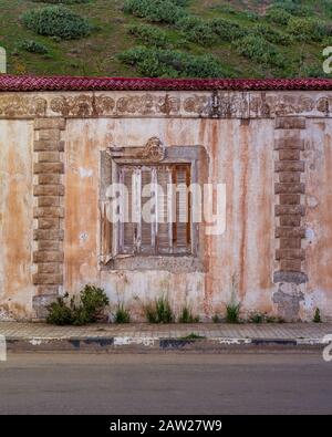 Exterior of aged house with weathered ornamental walls and shuttered window located under green hill and near asphalt road Stock Photo