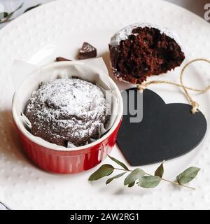Chocolate muffin in red cup. Mockup valentine black heart copyspace. Small glazed ceramic ramekin with brown cake on a white background Stock Photo