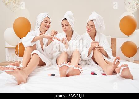 Front view of three smiling female friends in towels and bathrobes sitting on bed, doing pedicure and manicure. Cute happy young girls doing beauty procedures, enjoying time together, balloons nearby. Stock Photo
