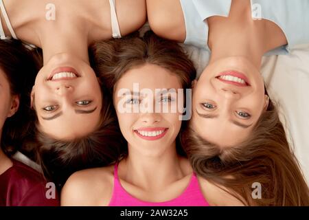 From above view of brunette young female friends lying on bed, posing and having fun. Close up of group of girls in casual outfits smiling, enoying time together at home. Concept of friendship. Stock Photo