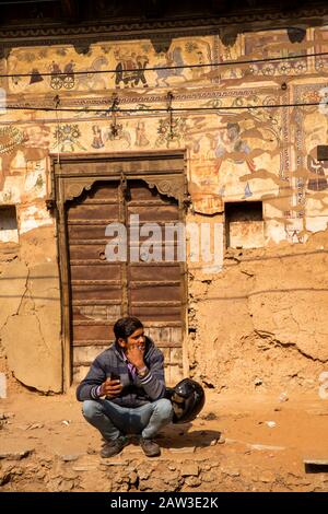 India, Rajasthan, Shekhawati, Mandawa, Akram Ka Haveli, man squatting below remnants of historic old painted walls with old wooden door Stock Photo
