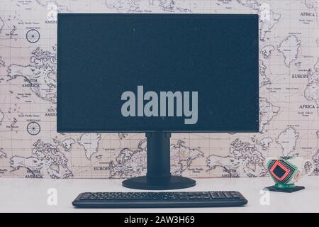 Blank computer desktop with keyboard and coffee mug on white table in sunny room, mock up. World map background. Black screen. Retro style look. Stock Photo