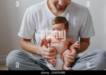 Happy Caucasian father holding newborn baby on laps knees. Man parent embracing rocking child daughter son. Authentic lifestyle candid moment. Proud y Stock Photo