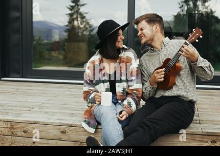 Hipster man playing on ukulele for his stylish woman, relaxing on wooden porch  of modern cabin with big windows in mountains. Happy young family trav Stock Photo